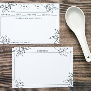 May include: Two white recipe cards with black leaf illustrations and text, including "RECIPE" and fields for ingredients and directions. A white ceramic spoon rests beside the cards on a wooden surface.