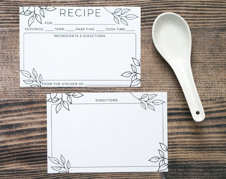 May include: Two white recipe cards with black leaf illustrations and text, including "RECIPE" and fields for ingredients and directions. A white ceramic spoon rests beside the cards on a wooden surface.