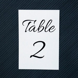 May include: A white table number card with the word "Table" in elegant black script above the number "2". The card is set against a dark blue textured background.