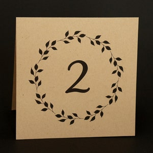 May include: A square, kraft paper table number card with the number "2" in a dark, elegant font. The number is encircled by a wreath of black leaves. The card is standing upright against a black background.
