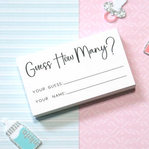 Op de afbeelding: Een stapel witte kaarten met de tekst "Guess How Many?" in zwart schrift. Daaronder staan lijnen voor "Your Guess:" en "Your Name:" op elke kaart. De achtergrond is een mix van lichtblauw en roze papier met patronen, met baby-thema versieringen.