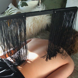 May include: A black fringe massage tool suspended above a person's back. The tool has a rectangular frame with long, thin black fringe hanging down. The person is lying on a bed with pillows in the background.