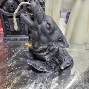 Peut inclure: Une bougie en forme de loup gris avec un design détaillé. Le loup hurle la gueule ouverte et les yeux fermés. La bougie est posée sur une surface grise.