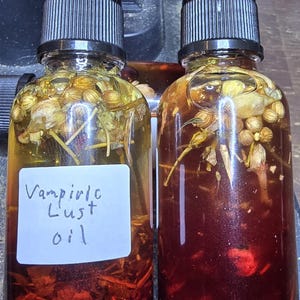 May include: Two glass bottles of oil with black droppers. One bottle is filled with a yellow liquid and the other with a red liquid. Both contain herbs and flowers. The label on one bottle reads "Vampiric Lust oil".