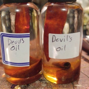 Puede incluir: Dos botellas de vidrio transparente con tapas negras, cada una con un líquido marrón rojizo y un pimiento rojo. Las etiquetas dicen "Devil's oil". Una botella también contiene una rama de canela. Las botellas están sobre una superficie de madera.