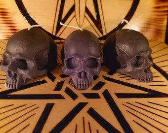 May include: Three black skull-shaped candles with white wicks. The candles are arranged in a triangular pattern on a wooden surface with a black and gold star design.