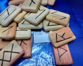 May include: A set of wooden runes with black markings on a light brown background. The runes are in a plastic bag with a label that says "Elder".