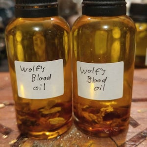 Könnte beinhalten: Zwei kleine bernsteinfarbene Glasflaschen mit schwarzen Schraubverschlüssen. Jede Flasche hat ein weißes Etikett mit schwarzer handschriftlicher Aufschrift "Wolf's Blood Oil".