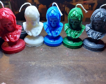 Può includere: Cinque candele a busto di teschio colorate. Le candele sono rosse, bianche, blu, verdi e grigie. Ogni candela ha uno stoppino e si trova su una base rotonda con la scritta "SANTA MUERTE" intorno.