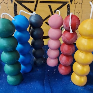 Puede incluir: Seis velas apiladas en forma de bola en varios colores: verde, azul, negro, rosa, rojo y amarillo. Cada vela tiene una mecha blanca en la parte superior. Las velas están dispuestas sobre una superficie azul con un fondo de madera.