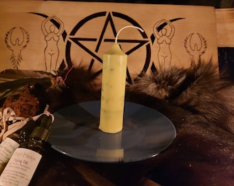 May include: A tall, pale yellow candle with green flecks stands on a blue plate. The candle is the focal point, with a wooden background featuring a pentagram and female figures. Small bottles of oil are visible in the foreground, suggesting a spiritual or metaphysical theme.