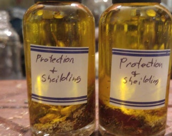 Peut inclure: Deux petites bouteilles en verre remplies d'un liquide jaune et d'herbes. Les bouteilles ont des bouchons noirs et des étiquettes blanches qui disent "Protection + Shielding".