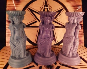 May include: Three decorative candles shaped like statues of a woman with three heads and a draped cloth. The candles are in shades of blue and purple.