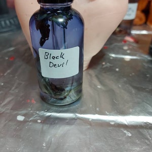 May include: A small glass bottle with a black cap filled with a blue liquid and dried flowers. The bottle has a white label that says "Black Devil".