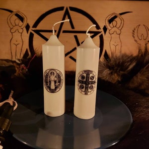 Może przedstawiać: Dwie białe świece z czarno-białymi obrazami religijnymi na niebieskim talerzu. Świece znajdują się na drewnianej powierzchni z brązowym futrem i małą butelką płynu.