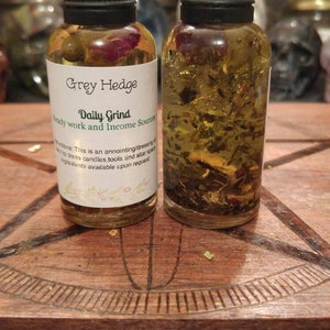 Könnte beinhalten: Zwei kleine Glasflaschen mit schwarzen Deckeln, gefüllt mit einer goldenen Flüssigkeit und Kräutern. Die Flaschen haben weiße Etiketten mit dem Text "Grey Hedge Daily Grind Ready work and Income Sources Instructions: This is an annointing/dressing to dress candles, tools and altar spaces. Ingredients available upon request."