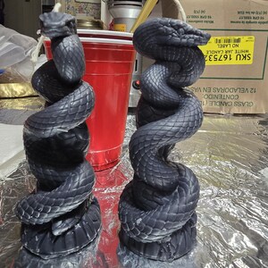 May include: Two black snake candles, each with a coiled body and a head with a forked tongue. The candles are on a silver surface.