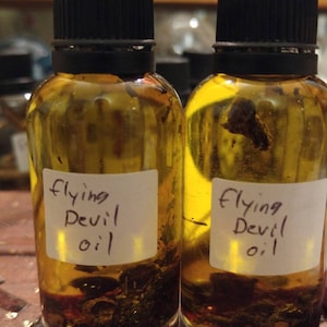 Puede incluir: Dos botellas de vidrio llenas de líquido amarillo y sedimento oscuro. Cada botella tiene una tapa negra y una etiqueta blanca con el texto manuscrito "Flying Devil oil". Las botellas están sobre una superficie de madera.