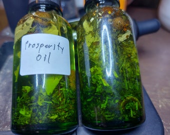 Peut inclure: Deux petites bouteilles en verre remplies d'un liquide vert et de diverses herbes et épices. La bouteille de gauche porte une étiquette blanche sur laquelle est inscrit "Prosperity Oil".