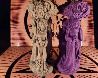 May include: Two decorative figurines of women with tree-like crowns. The figurine on the left is made of a light brown material and the figurine on the right is made of a purple material.