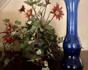 May include: A cobalt blue glass vase with a ruffled rim stands next to a floral arrangement. The vase has a bulbous base and a slender neck. The arrangement includes red and white flowers, green leaves, and trailing vines, all in a decorative pot.