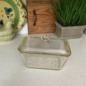 May include: A clear glass rectangular baking dish with a lid. The dish has a smooth, clear glass lid with a small knob on top. The dish is sitting on a white countertop.