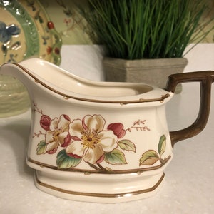 May include: A white ceramic gravy boat with a brown handle. The boat is decorated with a floral pattern of pink and yellow flowers with green leaves.