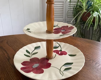 May include: Two-tiered serving stand with a wooden center post and cream-colored plates. The plates feature a floral design with burgundy flowers and green leaves. The stand is on a wooden table.