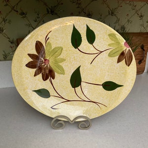 May include: A yellow ceramic plate with a brown and green floral design. The plate has a speckled glaze and is decorated with stylized leaves and flowers.