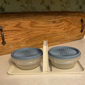 May include: A set of two small, clear plastic bowls with light blue lids, sitting on a rectangular tray. The lids have a radial design. A white divider separates the bowls. A wooden serving tray is in the background.