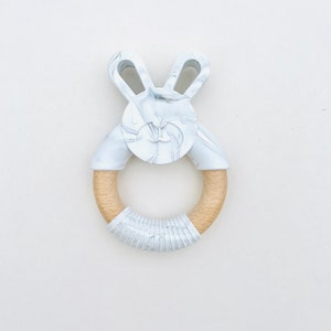 Puede incluir: Un mordedor para bebé con forma de conejito, con un diseño veteado blanco y gris. Las orejas y la cara del conejito están conectadas a un anillo de madera, envuelto en tela blanca. Un producto para bebés.