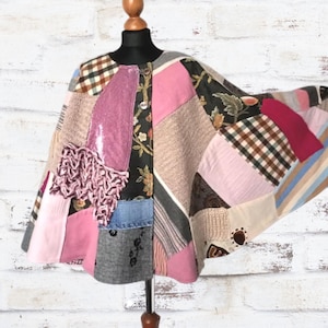 May include: A patchwork cape with a variety of fabrics, including pink, brown, and black floral patterns. The cape is displayed on a mannequin against a white brick wall. The design includes squares and rectangles of different textures.