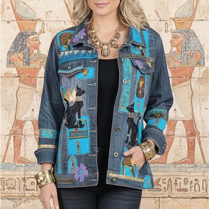 Könnte beinhalten: Jeansjacke mit ägyptischen Motiven. Die Jacke hat blaue Paneele mit Hieroglyphen, Illustrationen schwarzer Katzen und lila Blumenakzenten. Der Hintergrund zeigt altägyptische Wandkunst.