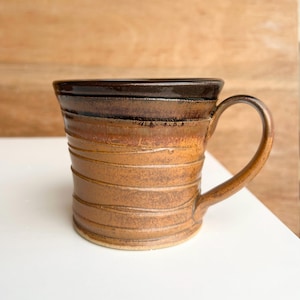 May include: A handmade ceramic mug with a brown and tan colour gradient. The mug features a textured, ridged design around the body and a curved handle. The top rim is a darker brown, and the mug appears to be glazed.
