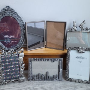 May include: A collection of silver-toned picture frames with various designs. The frames are arranged on a wooden surface. The frames feature different designs, including a birthday theme, a Chicago skyline, and a simple design with a bow and bells. The frames are all made of metal and have a vintage look.