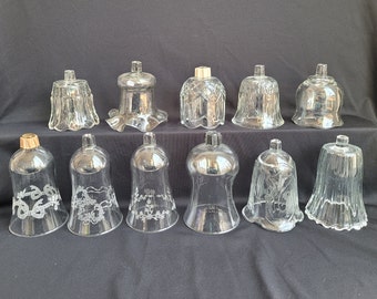 May include: A collection of clear glass candle holders in various shapes and designs. Some have etched floral or ribbon patterns, while others feature textured or fluted surfaces. The candle holders are displayed on a dark background.