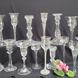 May include: A collection of clear glass candle holders in various designs. The holders range in height and style, including fluted, twisted, and flared shapes. Some have intricate cut glass patterns, while others are smooth. A few pink flowers are in the foreground.