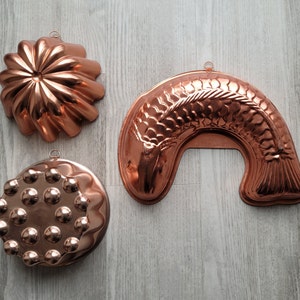 May include: Three copper-colored cake molds. One is shaped like a flower, one is shaped like a circle with raised dots, and one is shaped like a fish.