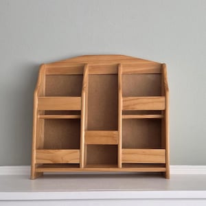 May include: A wooden wall-mounted organizer with a light brown finish. It features three sections: a central area and two side compartments, each with a shelf. The top has a curved design.