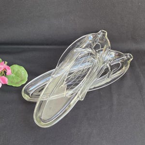 May include: Two clear glass serving dishes with an elongated shape, resting on a dark surface. The dishes have a decorative design and are positioned near a pink flower with green leaves. The dishes are likely for serving appetizers or small portions.