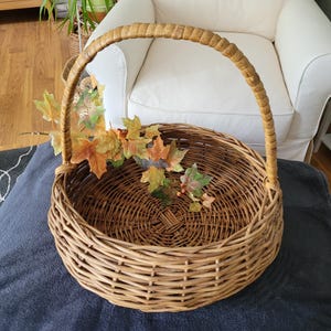 May include: A large, round wicker basket with a woven handle. The basket is empty and has a natural brown color.  A sprig of fall leaves with red berries is inside the basket.