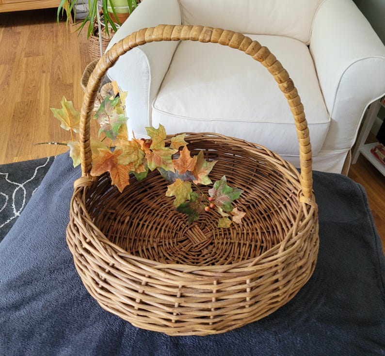 May include: A large, round wicker basket with a woven handle. The basket is empty and has a natural brown color.  A sprig of fall leaves with red berries is inside the basket.