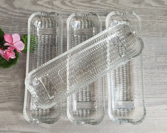 Puede incluir: Un conjunto de fuentes de vidrio transparente. Cada fuente rectangular tiene una superficie texturizada y extremos redondeados. Las fuentes están dispuestas sobre una superficie de madera clara, con una flor rosa en el fondo.
