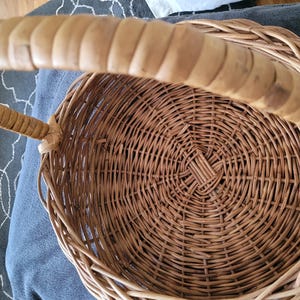 May include: A woven wicker basket with a natural brown finish and a long, curved handle. The basket is empty and has a circular shape.