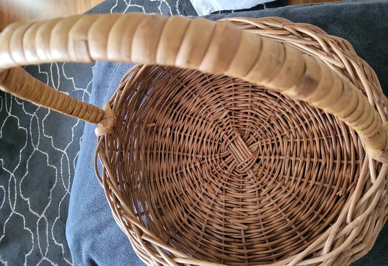 May include: A woven wicker basket with a natural brown finish and a long, curved handle. The basket is empty and has a circular shape.