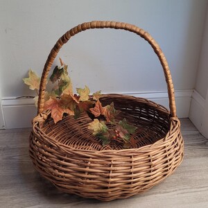 May include: A large, round wicker basket with a single, curved handle. The basket is made of natural brown wicker and has a woven pattern. The basket is empty, but there are some fall leaves inside.