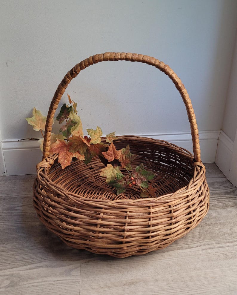 May include: A large, round wicker basket with a single, curved handle. The basket is made of natural brown wicker and has a woven pattern. The basket is empty, but there are some fall leaves inside.