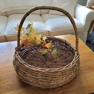 May include: A large, round wicker basket with a woven handle. The basket is filled with artificial fall leaves in shades of green, yellow, and orange.