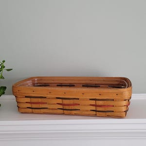 May include: A rectangular woven wood basket with a light tan finish, featuring horizontal bands of dark green and red accent strips woven through the sides. The basket has a flat rim and a sturdy construction, showcasing a classic woven design.