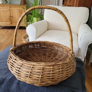 May include: A large, round, woven wicker basket with a brown handle. The basket is empty and sitting on a gray blanket.